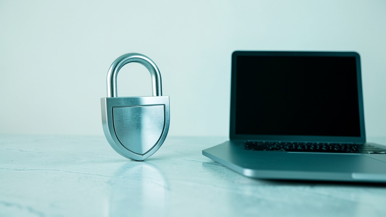 A padlock icon with a shield, placed on a desk next to a laptop, representing privacy and security.